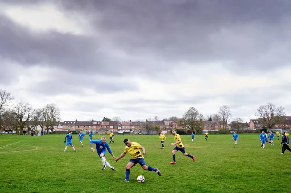 An amateur match on a park pitch, with a player in yellow taking the ball past a defender in blue under a grey sky.